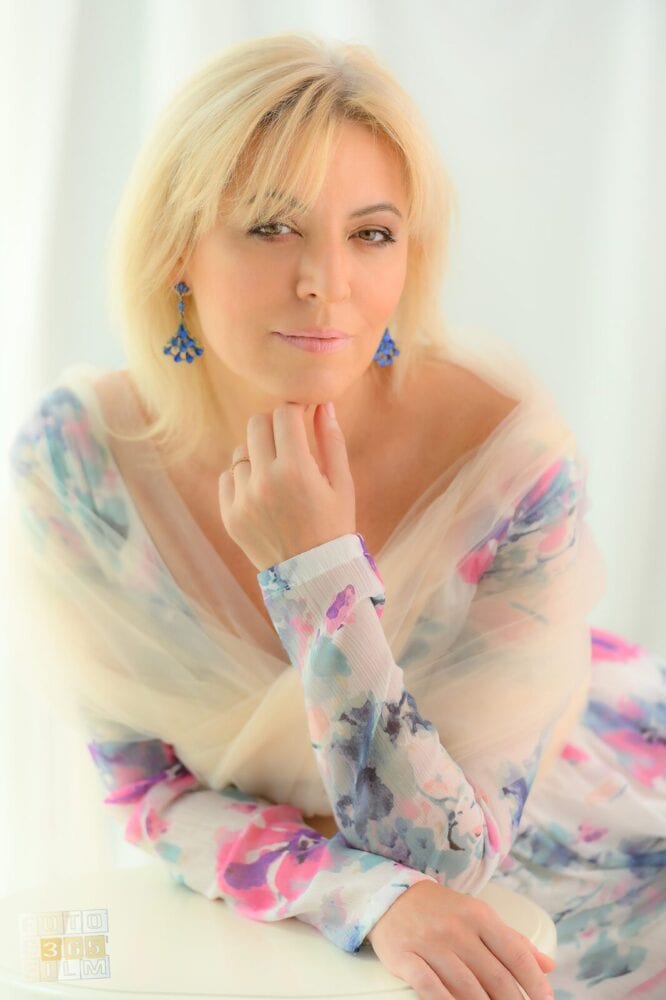 Fotografie facuta la o sedinta foto glamour pentru femei, reprezentand o femeie blonda cu rochie crem cu imprimeuri, cercei albastri, pe fundal alb